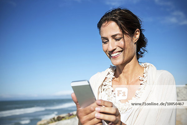 Südafrika  Porträt einer lächelnden Frau mit Blick auf das Smartphone vor dem Meer