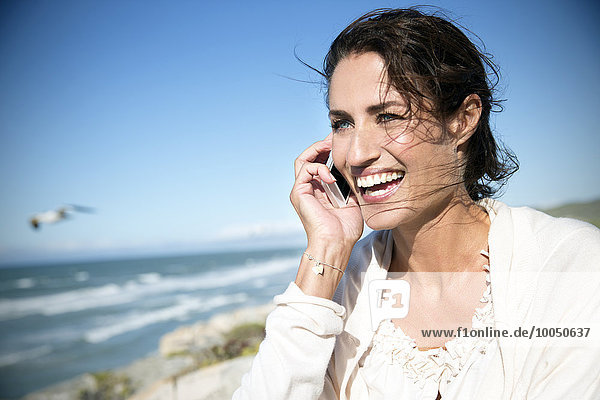Südafrika  Porträt einer lächelnden Frau beim Telefonieren mit dem Smartphone vor dem Meer