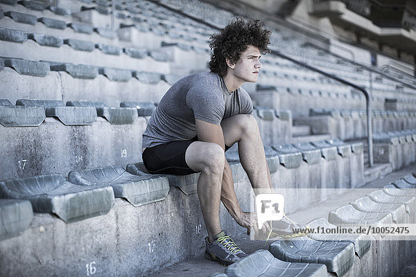Junger Sportler sitzt auf der Tribüne eines Stadions und bindet seine Schuhe.