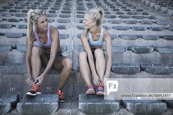 Zwei Sportlerinnen sitzen auf der Tribüne eines Stadions und binden ihre Schuhe.