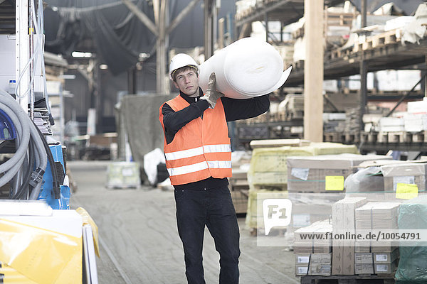 Young male warehouse worker carrying polystyrene roll on shoulder