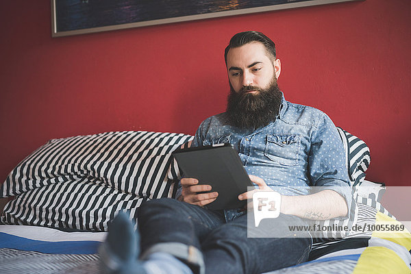 Junger bärtiger Mann mit digitalem Tablett auf dem Bett