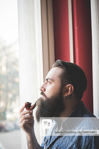 Young bearded man smoking pipe by window