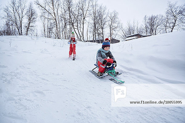 Zwei Brüder auf Schlitten und Schneescooter auf schneebedecktem Hügel  Hemavan  Schweden