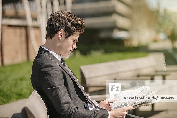 Ein junger Geschäftsmann sitzt auf einer Bank und liest Zeitung.