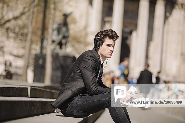 Young city businessman with newspaper sitting on bench listening to headphones