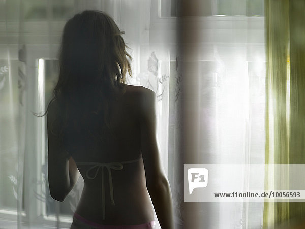 Silhouettierte Rückansicht einer jungen Frau  die aus dem Fenster schaut.