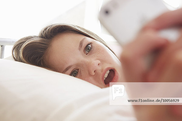 Überraschtes Mädchen liegt auf dem Bett und liest eine Smartphone-Nachricht.