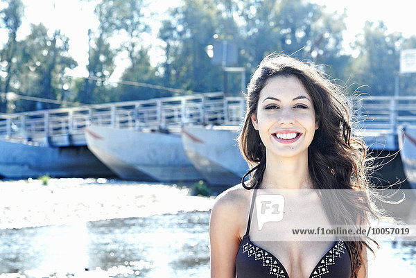Porträt einer lächelnden jungen Frau im Bikini vor einem Fluss stehend