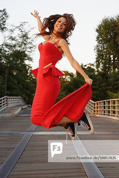 Porträt einer jungen Frau in einem schicken roten Kleid und Baseballschuhen  die in Bewegung springen.