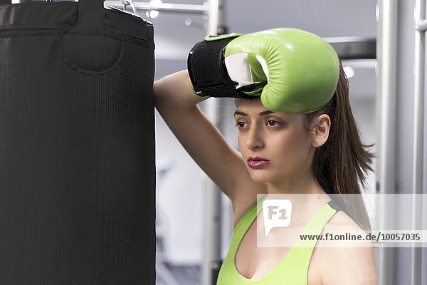 Portrait einer jungen Frau im Fitnessstudio  die sich gegen einen Boxsack mit Boxhandschuh lehnt.