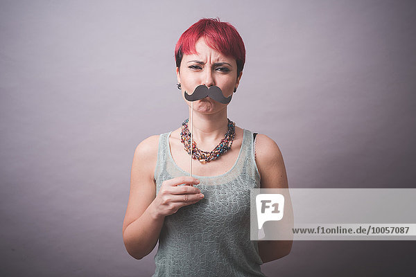 Atelierporträt einer verwirrten jungen Frau  die den Schnurrbart vor dem Gesicht hält.