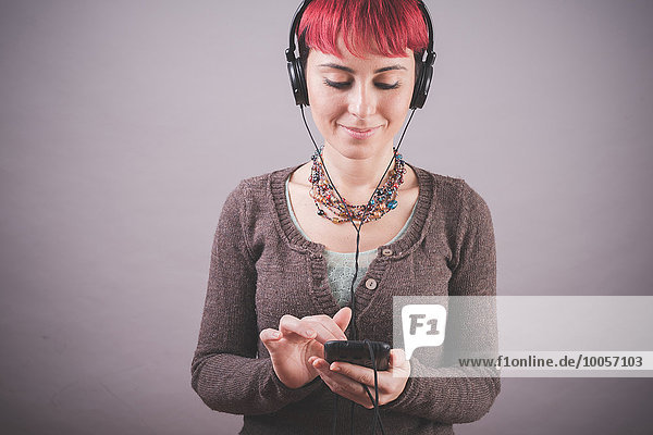 Studio-Porträt einer jungen Frau mit kurzen rosa Haaren bei der Musikauswahl auf dem Smartphone