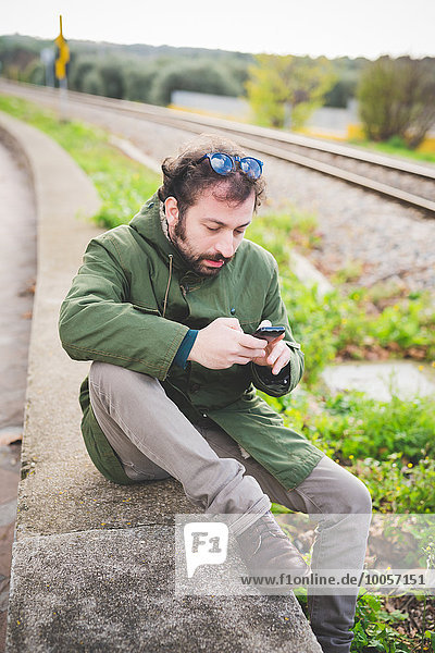 Mid adult man sitting on wall next to railway track using smartphone