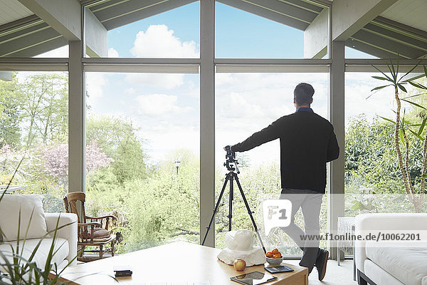 Rückansicht eines Mannes  der sich auf ein Teleskop stützt und aus dem Fenster blickt und die Gartenlandschaft bewundert.