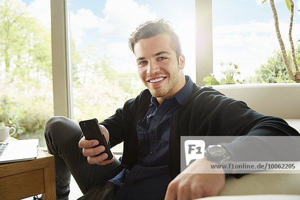 Porträt eines lächelnden Mannes,  der auf dem Boden sitzt und ein Smartphone hält.