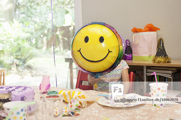 Mädchen sitzend am Geburtstagstisch mit Kuchenspiel mit Smiley-Gesichtsballon