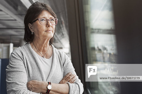 Thoughtful senior businesswoman standing arms crossed by window in office