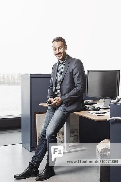 Full length portrait of confident businessman leaning on desk in office