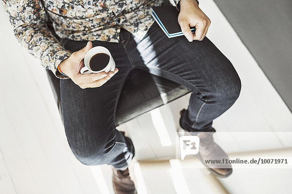 High angle view of man holding coffee cup and note pad at home office