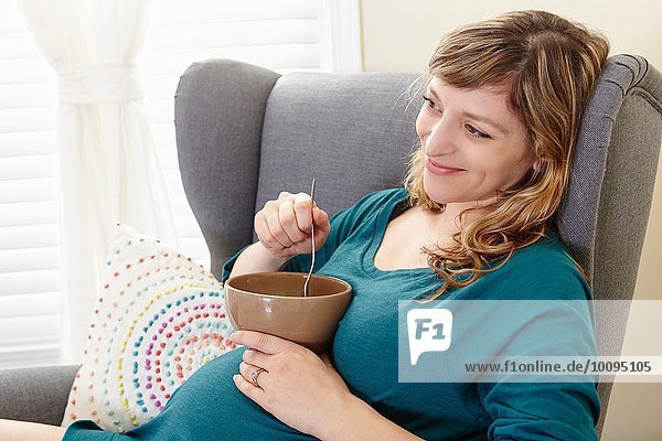 Schwangere Frau isst Suppentasse auf Wohnzimmersessel
