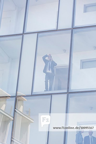 Geschäftsmann schaut durch das Fenster eines Hotels mit Glasfront  während er auf dem Smartphone spricht.
