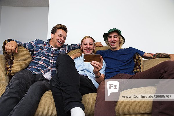 Three young men sitting on sofa  looking at smartphone