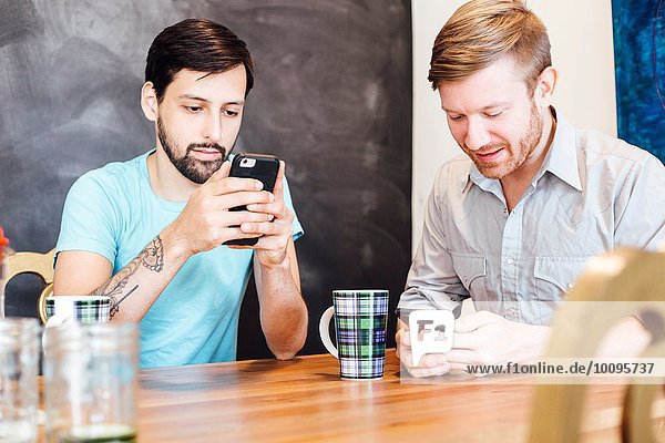Männliches Paar am Tisch sitzend  Kaffee trinkend  Smartphones anschauend
