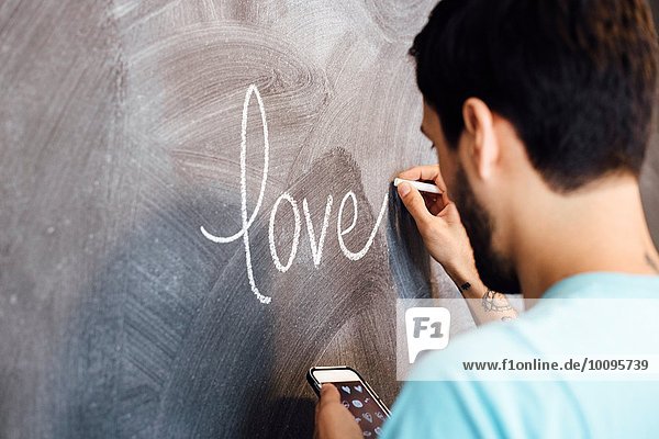 Junger Mann  das Wort Liebe auf die Tafel schreiben  Smartphone halten  Rückansicht