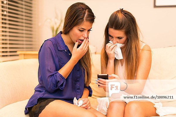 Two young women  sitting on sofa looking at smartphone  shocked expression