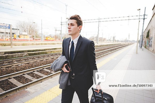 Young businessman commuter walking along railway platform with suitcase.