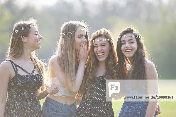 Vier Teenager-Mädchen in Gänseblümchen-Kopfbedeckungen kichern im Park.