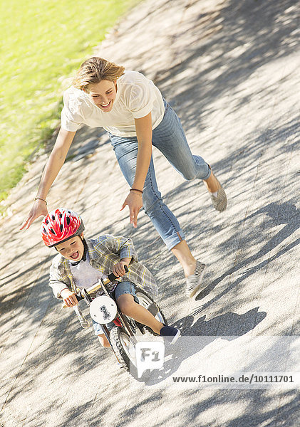 Mother pushing son with helmet on bicycle in sunny park