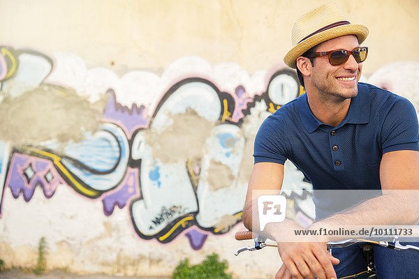 Smiling man in hat and sunglasses leaning on bicycle next to urban graffiti wall