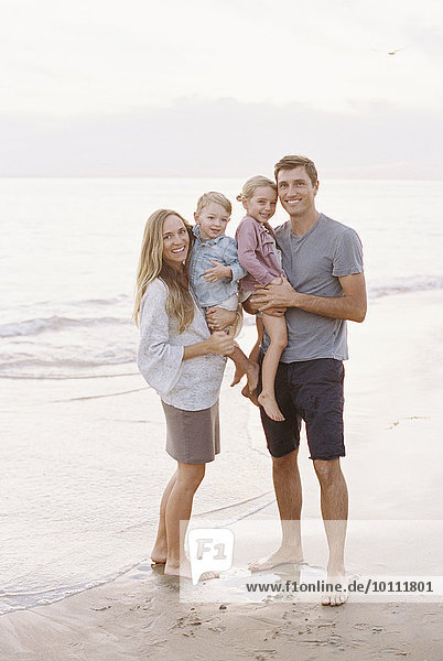 Couple standing with their son and daughter on a sandy beach by the ocean  looking at camera  smiling.