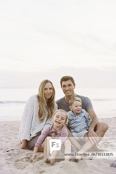 Couple sitting with their son and daughter on a sandy beach by the ocean  looking at camera  smiling.