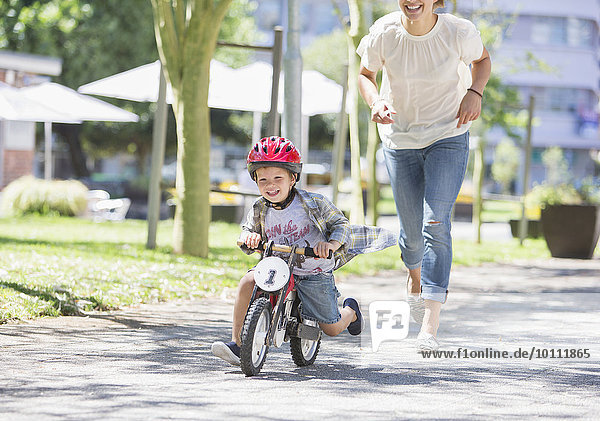 Mother chasing son riding bicycle with helmet in sunny park