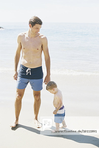 Man and his son wearing swimming trunks standing on a sandy beach by the ocean.