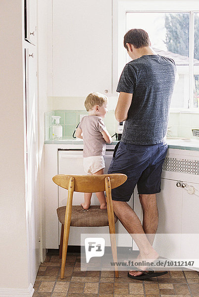 Man standing in a kitchen  his son standing on a chair beside him.