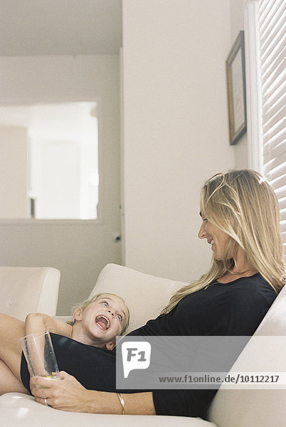 Woman with long blond hair wearing a black dress  sitting on a sofa  holding a glass  playing with her daughter.