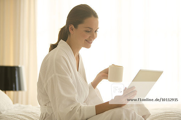 Woman in bathrobe drinking coffee and using digital tablet in bedroom