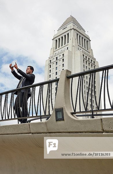 Geschäftsmann  der auf dem Gehweg steht und mit einem digitalen Tablett fotografiert  Los Angeles City Hall  Kalifornien  USA