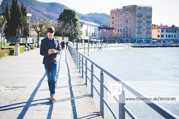 Junger Mann beim Bummeln und Lesen von Smartphone-Texten am Seeufer  Rovato  Brescia  Italien