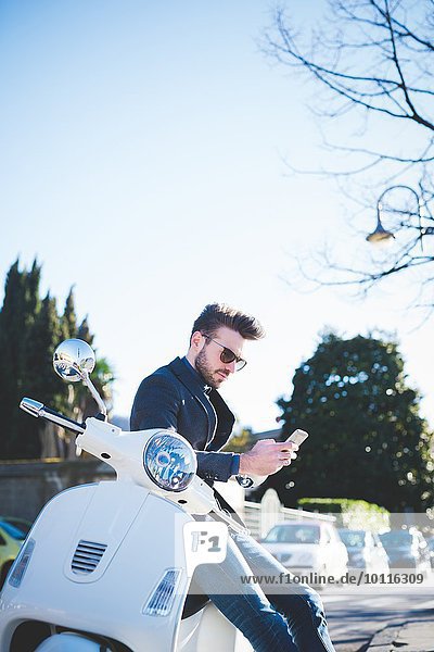 Stilvoller junger Mann beim Lesen von Smartphone-Texten auf Moped