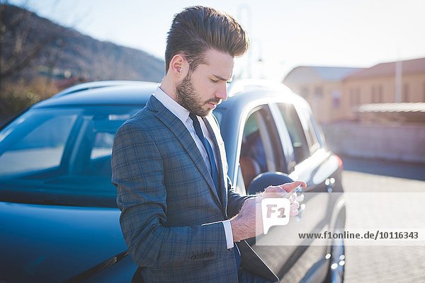 Junger Geschäftsmann  der sich gegen das Auto lehnt und Smartphone-Texte liest.