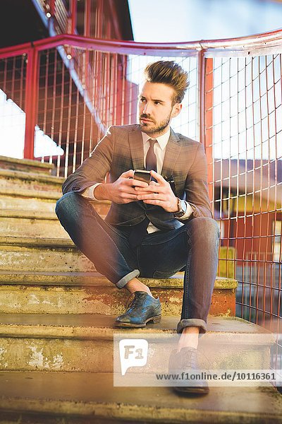 Stilvoller junger Mann mit Smartphone auf Stegstreppe sitzend