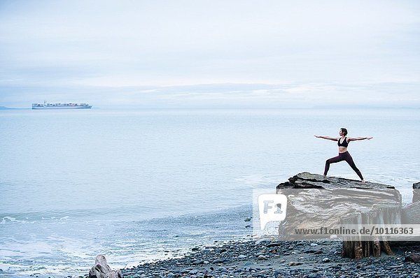 Reife Frau  die Yoga-Pose übt  während sie auf einem großen Treibholz-Baumstumpf am Strand steht.