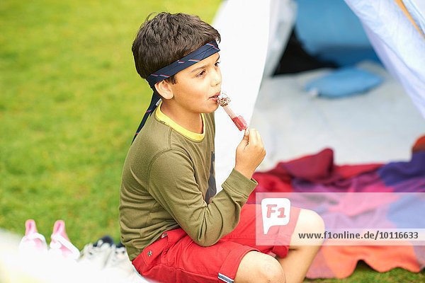 Junge isst Eis am Stiel vor hausgemachtem Zelt im Garten