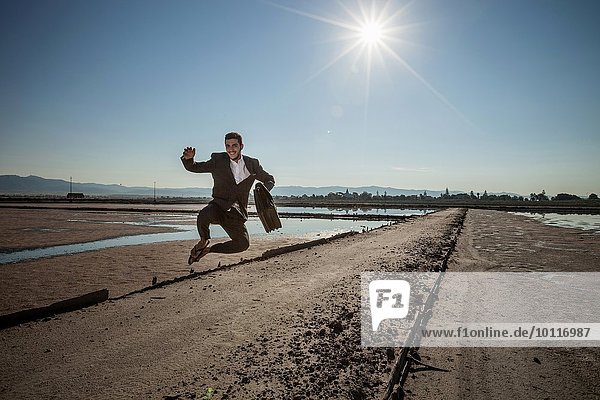 Mid adult businessman carrying briefcase jumping mid air at beach