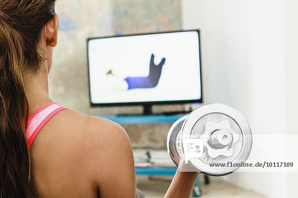 Junge Frau,  die mit Handgewichten im Wohnzimmer trainiert,  während sie den Computerbildschirm beobachtet.